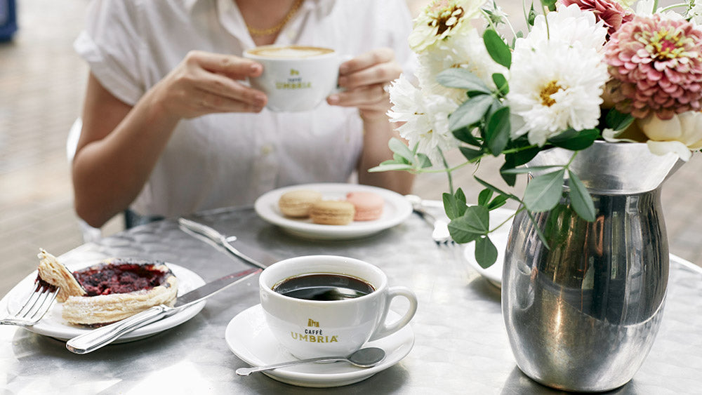 A person in a white shirt holds a white coffee cup with gold lettering at a table. In the foreground is another cup of black coffee on a saucer, a pastry on a plate, and a large silver vase filled with white and pink flowers.