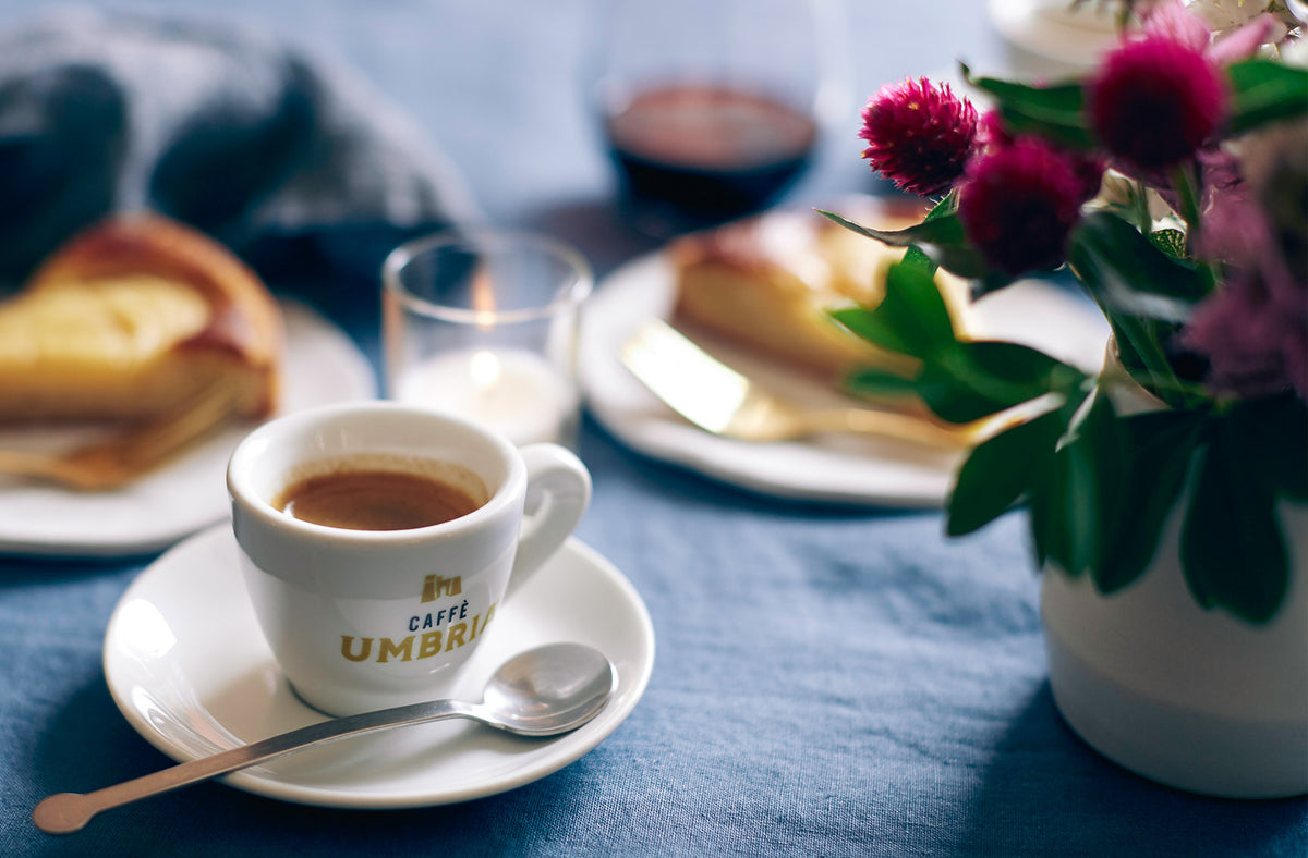 A white ceramic espresso cup with 'CAFE UMBRIA' printed in gold on the side, filled with espresso and resting on a matching saucer with a spoon. The scene is styled with a blue tablecloth, a slice of cake, a glass of milk, and a vase of pink flowers in the background.