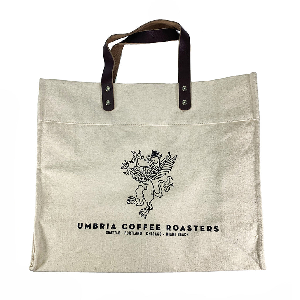 A beige cotton tote bag with leather shoulder straps and a logo printed on the front.